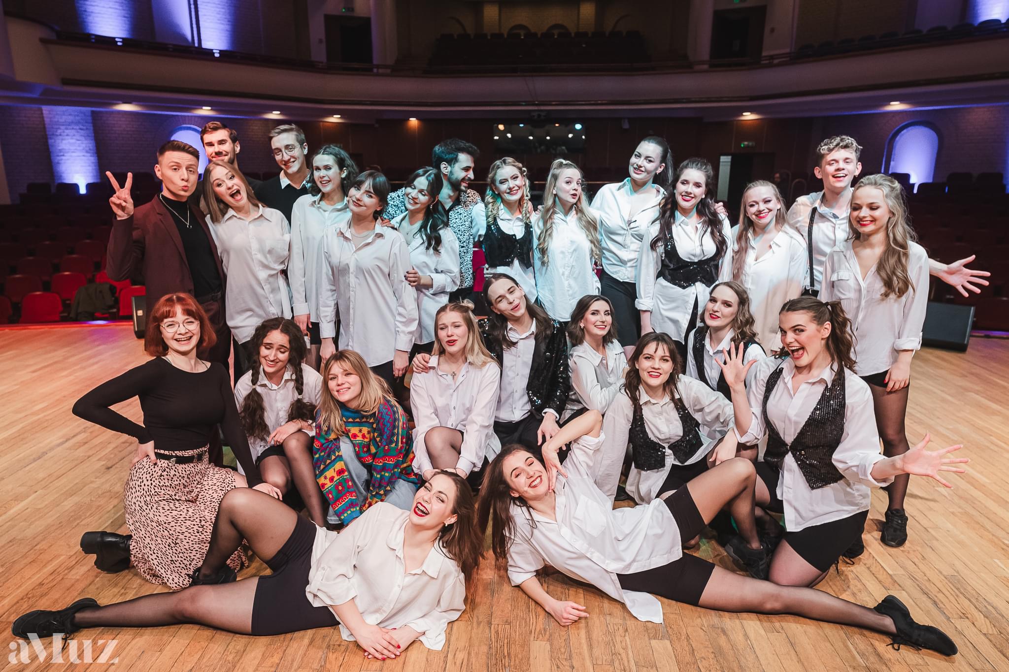 Duża grupa młodych os&oacute;b pozuje wsp&oacute;lnie na scenie sali koncertowej, uśmiechnięci i&nbsp;w swobodnych, scenicznych stylizacjach.