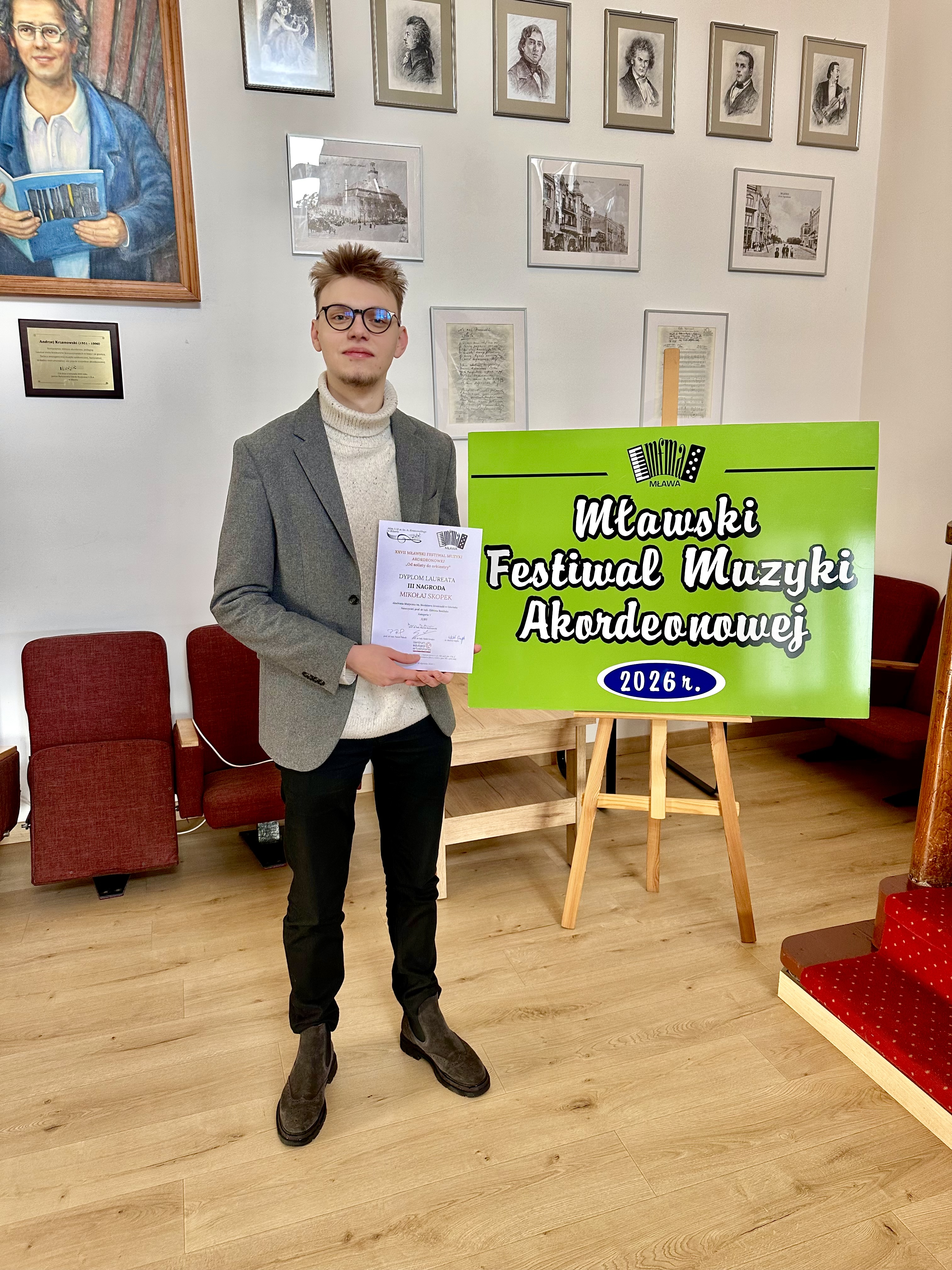 Zdjęcie przedstawiające Mikołaja Skopka, kt&oacute;ry trzyma w&nbsp;ręce dyplom. Z&nbsp;tyłu tablica z&nbsp;namisem "Mławski Festiwal Muzyki Akordeonowej".